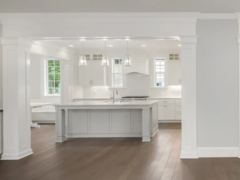 An open-plan white kitchen seen through a columned opening, with a painted island, glass pendants and wide plank floors running through to the next room